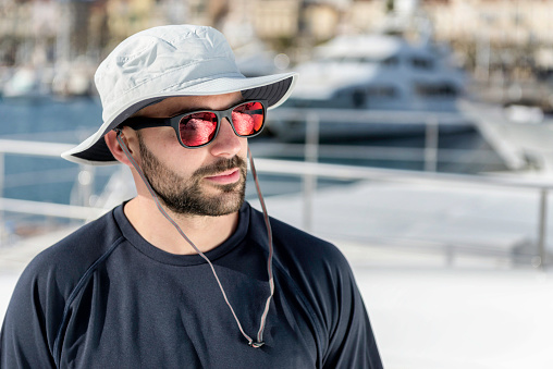 Horizontal color portrait image of beautiful male fashion model posing on luxury yacht port of Cannes, France, brown-haired male wearing cap and reflective sunglasses. Focus on attractive male model, luxury blurred yacht in background.
