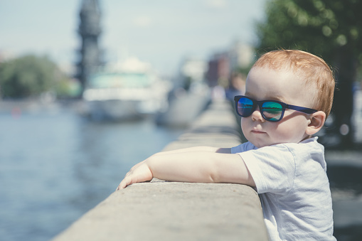 Portrait of a cute red-haired boy in sunglasses, holding on to the stone parapet of the waterfront park