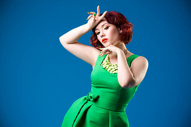 Young red-haired woman in green dress posing on blue background Young beautiful red-haired caucasian woman in green dress posing in studio on blue background, professional makeup and hairstyle, expressive portrait fine art portrait pin up girl glamour beauty stock pictures, royalty-free photos & images