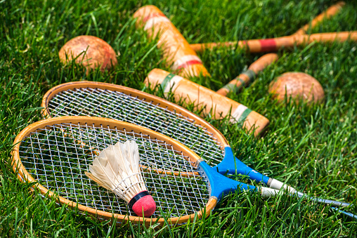 A pair of vintage wooden badminton rackets and a feathered shuttlecock lying in grass, along with a pair of croquet mallets and balls in the background. Great old fashion family fun games for all ages.