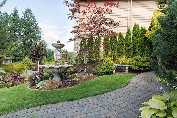 House manicured frontyard garden with water fountain stone bench green lawn plants trees shrubs and brick paver walkway path