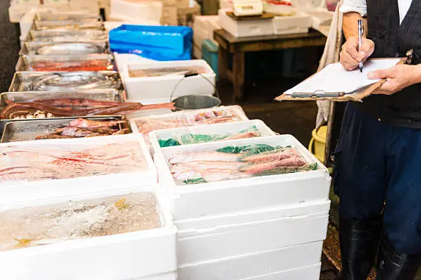Tsukiji, fishmarket in Tokyo is a great turistic sight not just large commercial activity.  On a tour you can visit the world's largest fish market. There is a labyrinth of seafood stalls, youn can try to make your own sushi. Here visible part of man taking notes on a pad standing beside seafood in boxes with ice. Japan. 