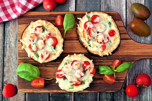 Healthy eggplant mini pizzas with melted mozzarella, tomatoes and basil on a serving board against rustic wood