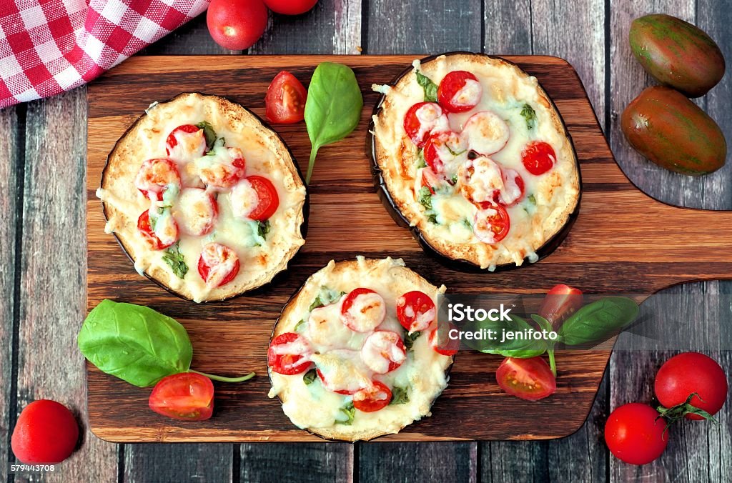 Healthy eggplant pizzas with melted mozzarella, tomatoes and basil Healthy eggplant mini pizzas with melted mozzarella, tomatoes and basil on a serving board against rustic wood Eggplant Stock Photo