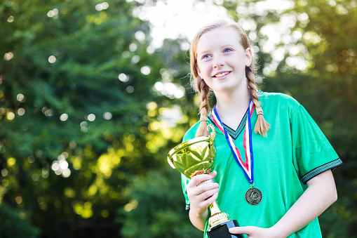 Beautiful caucasian girl with strawberry blonde hair. She is wearing a green soccer uniform and a winners medal around her neck. She is holding the teams winning soccer trophy. She has braids in her strawberry blonde hair. She is smiling at someone/something off camera. She is standing in front of a tree, the sun is coming in through the branches.