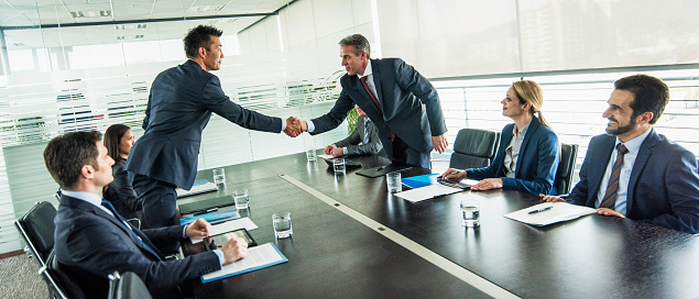 Two businessmen standing and shaking hands across a conference table. Other people sitting around the table.