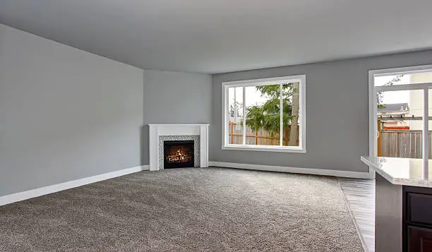 Grey house interior of living room with fireplace and carpet floor. Windows overlooking back yard.
