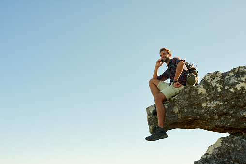 Shot of a hiker talking on his phone while sitting on the edge of a cliff