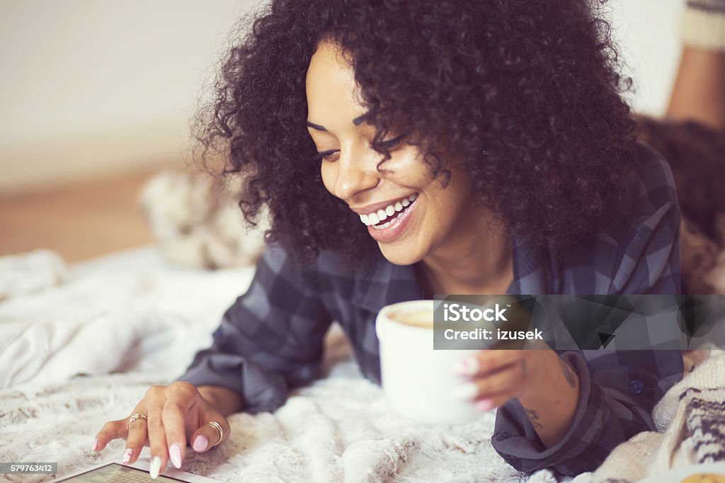 Afro american woman using a digital tablet at home Beautiful afro american young woman lying on the stomach on a bed and using a digital tablet and drinking coffee. Winter scenery, close up of face. Winter Stock Photo