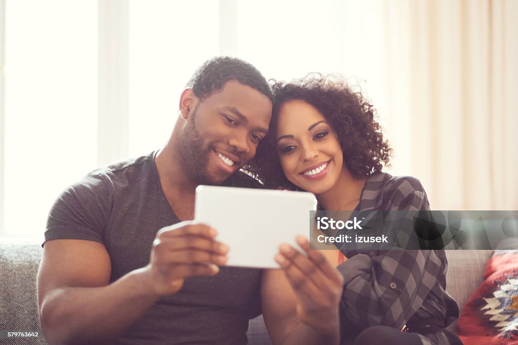 Afro american couple using a digital tablet at home Afro american friendly couple sitting on sofa at home, taking selfie using a digital tablet. Adult Stock Photo