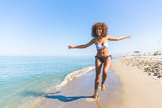 Young black woman having fun at seaside Young black woman having fun at seaside. She is twenty years old, mixed race caucasian and african black, with curly and voluminous hair, running with open arms and happy face. one piece swimsuit photos stock pictures, royalty-free photos & images