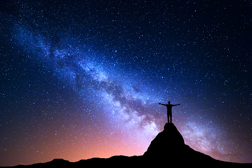 Night landscape with Milky Way. Silhouette of a standing man with raised up arms on the mountain peak . Beautiful Universe. Travel background with blue night starry sky and city lights