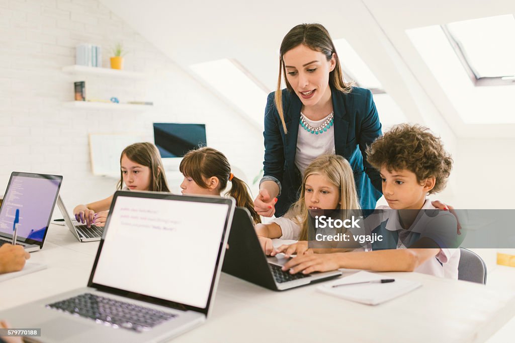 Kids Coding In School Group of children at coding class. They are sitting by the desk and using laptops. Their female teacher standing beside them and helping with their assignment. Child Stock Photo