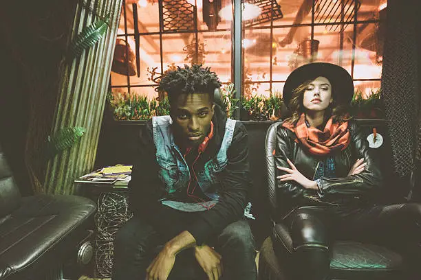 Fashion portrait of contemporary afro american guy and beautiful woman sitting in cool pub and looking at camera.