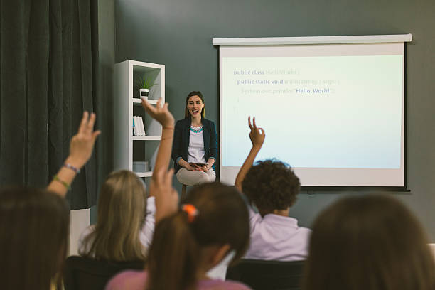 Kids Coding In School. Group of children at coding class. Teacher sitting and asking questions and children raising hands to answering. teacher projector stock pictures, royalty-free photos & images