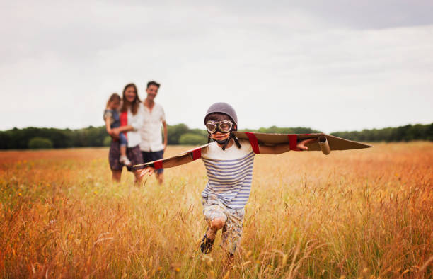 Playful boy with wings in aviators cap and flying goggles in field  21st century stock pictures, royalty-free photos & images