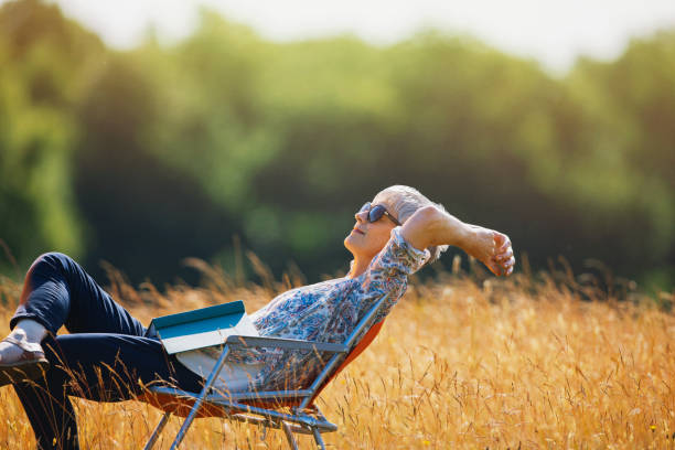 mujer mayor despreocupada relajándose con un libro en un campo soleado - women book mature adult reading fotografías e imágenes de stock