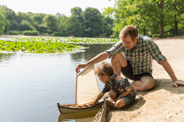 vater und sohn spielen mit spielzeugsegelboot am seeufer - sailing nautical vessel family lake stock-fotos und bilder