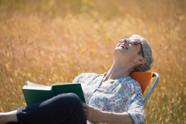 mujer mayor leyendo libros y riendo con la cabeza hacia atrás en un campo soleado - women book mature adult reading fotografías e imágenes de stock