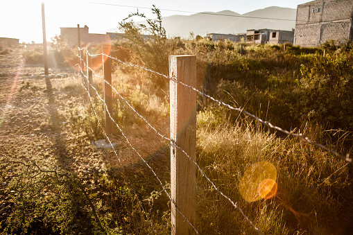 Diminishing perspective view down a barbed wire fence in the Mexican town of Saltillo.  Impoverished buildings, homes.  Lens flare.  No people.
