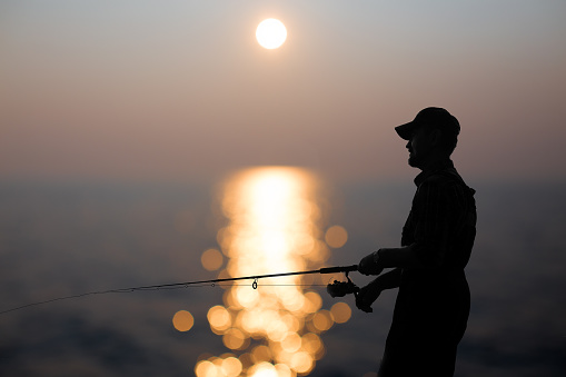 silhouette of a fisherman with a fishing rod in the background reflected in water sunset. blur sunlight on the water