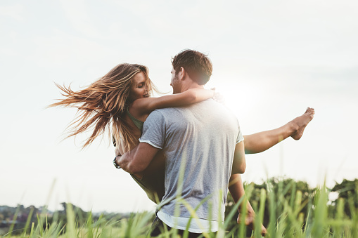 Shot of young man carrying his girlfriend in his arms on grass field. Couple having fun in nature on a summer day.