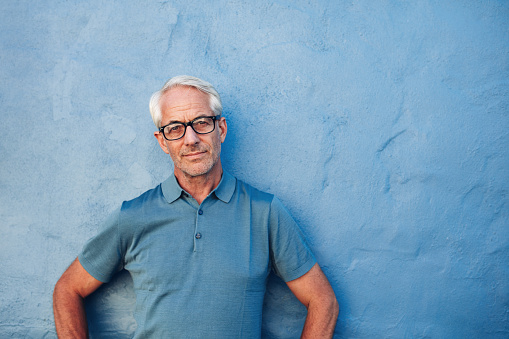 Portrait of a mature man standing against a blue background with copy space. Caucasian man wearing glasses leaning to a wall and staring at camera.