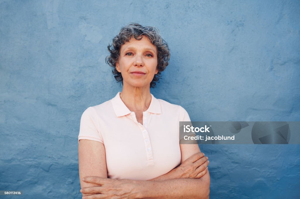 Confident mature woman standing with her arms crossed Portrait of confident mature woman standing with her arms crossed against blue background Serious Stock Photo