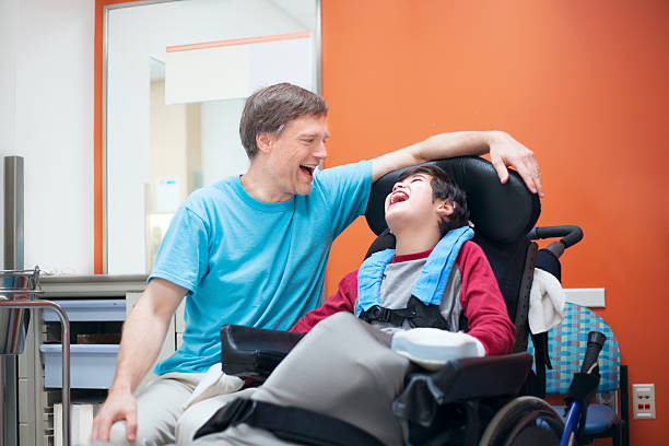 Disabled little boy in wheelchair talking with father in hospital room Father talking with disabled biracial son sitting in wheelchair while waiting in doctor's office, laughing together. developmental disability stock pictures, royalty-free photos & images