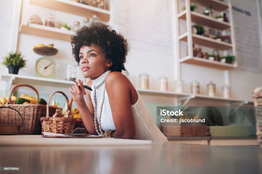 Young african female juice bar owner Indoor shot of young african female juice bar owner looking away and thinking, she is standing behind bar counter. Contemplation Stock Photo