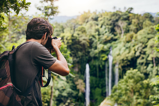 Man with backpack standing in front of waterfall and taking a photo. Male hiker photographing a beautiful water fall in forest