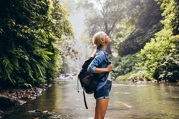 Relaxed female hiker standing by the stream Side view shot relaxed young woman with backpack standing by the stream. Female hiking by the creek. eco tourism photos stock pictures, royalty-free photos & images