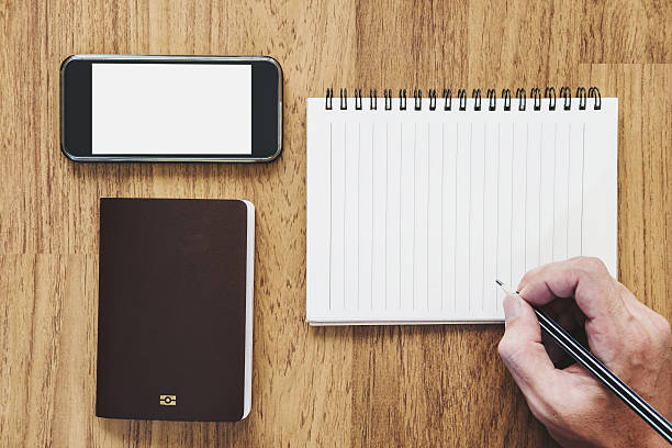 Hand holding pencil writing notebook and mobile phone with passport on wooden desk, vintage tone
