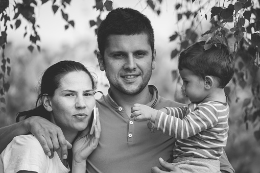 Monochrome photo of smiling caucasian family of father, mother, and cute daughter posing in embrace at studio. Happy parents with female toddler expressing pure love and sincere feelings.