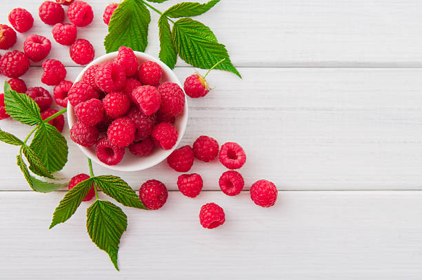 Red fresh raspberries on white rustic wood background Red fresh raspberries on white rustic wood background. Bowl with natural ripe organic berries with peduncles and green leaves on wooden table, top view with copy space raspberry photos stock pictures, royalty-free photos & images