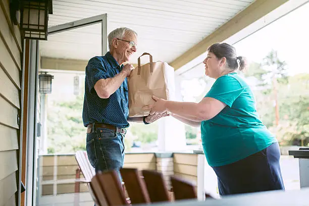 A kind and caring neighbor or friend delivers fresh produce from the grocery store to an elderly man at his home.  He receives the gift with a smile on his face, grateful for the help and assistance.  Horizontal image.