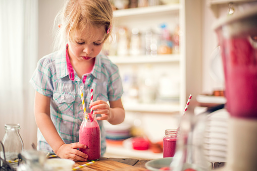 Two little girls drinking fresh smoothie at home. They are making smoothie with fresh fruits, milk and some juice in a electric mixer.