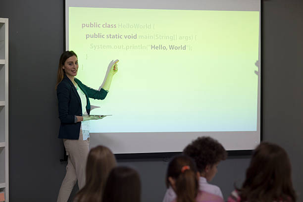 Kids Coding In School. Group of children at coding class. Teacher standing, holding digital tablet and having presentation. Children sitting in front of her and listening carefully. Teacher is talking about coding. teacher projector stock pictures, royalty-free photos & images