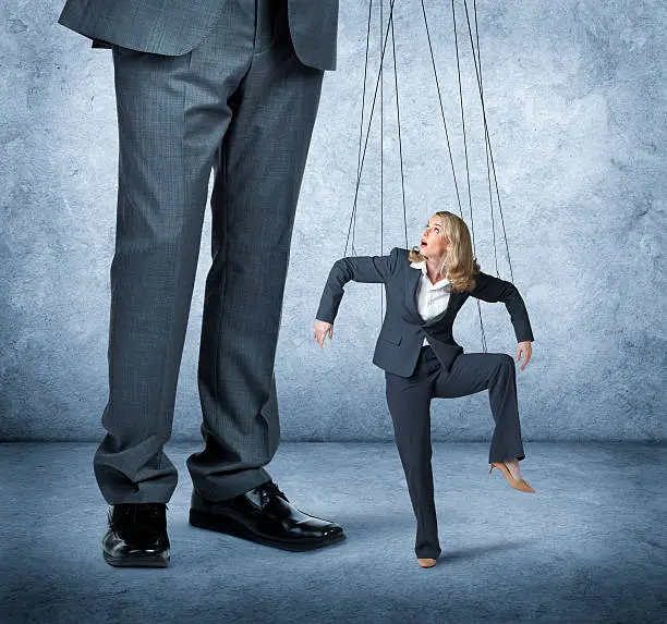 A large businessman controls the movement of a businesswoman as she is controlled by the strings of a marionette.