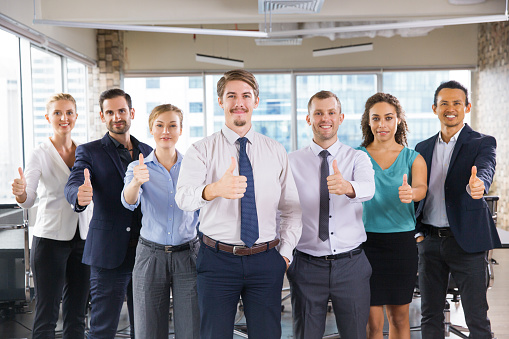 Group of successful young business people standing in office, showing thumbs up sign and smiling. Business success concept