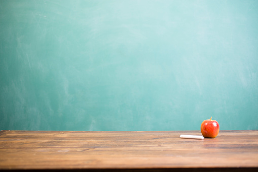 It's back to school time!  A red apple with piece of white teacher's chalk.   The objects lie on top of a wooden school desk with a green chalkboard in far background.  The blank blackboard in the background makes perfect copyspace!  Education background themes.