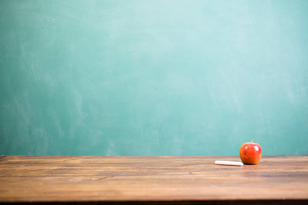 Red apple with chalk and school chalkboard. It's back to school time!  A red apple with piece of white teacher's chalk.   The objects lie on top of a wooden school desk with a green chalkboard in far background.  The blank blackboard in the background makes perfect copyspace!  Education background themes. school board stock pictures, royalty-free photos & images