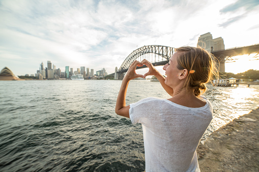 Young woman in Sydney harbour makes a heart shape finger frame on the Sydney skyline.