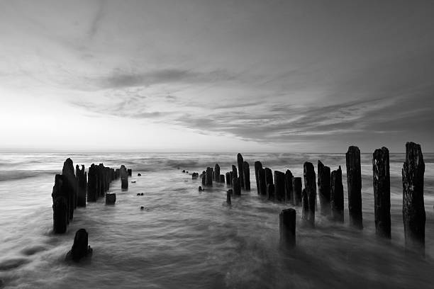 Beach poles in the sea on a cloudy day. Black and white image with a long exposure creating a smooth water surface. The poles are in line to a vanishing point on the horizon over the water.