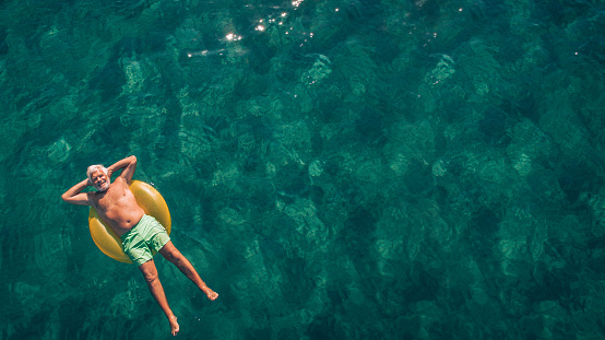 High angle view photo of a senior man relaxing while floating in the ocean using swimming tube; wide photo dimensions