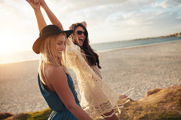 Female friends enjoying a day at sea coast Shot of stylish young women enjoying at the sea coast. Female friends walking together at the beach on a summer day and having fun. beach fashion stock pictures, royalty-free photos & images