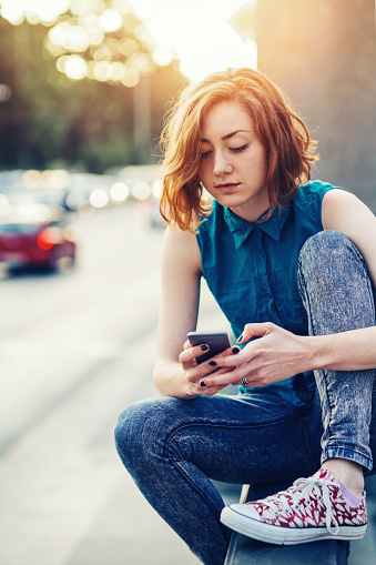 Young girl texting on a mobile phone and waiting for her boyfriend while sitting on a boulevard bridge downtown