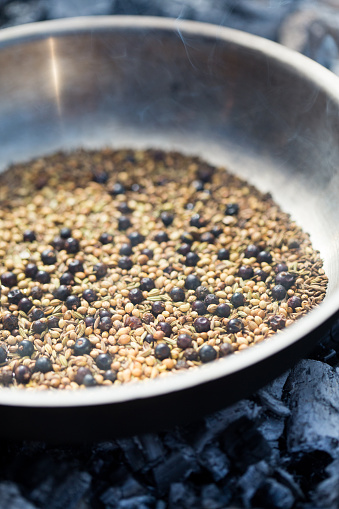 A frying pan full of dry spices including juniper berries, black pepper, fennel, cumin and coriander, being dry roasted on a bed of hot embers.