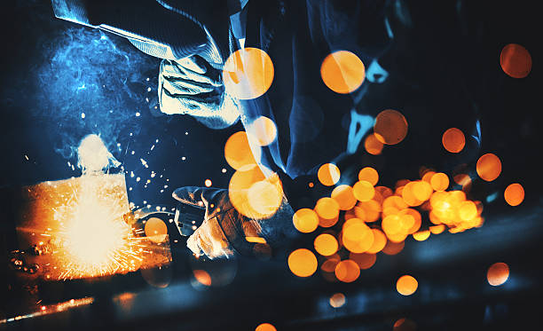 Welding at a workshop. Closeup side view of two workers processing steel pieces by grinding and welding. Welder is in sharp focus, using arc welding machine. Toned image. steel grinding stock pictures, royalty-free photos & images