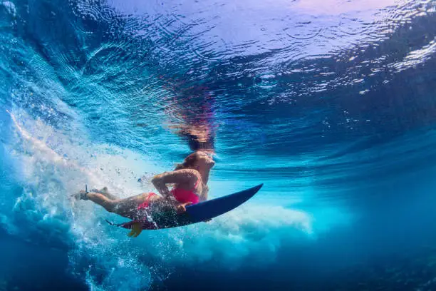 Young active girl wearing bikini in action - surfer with surf board dive underwater under big ocean wave. Family lifestyle, people water sport adventure camp and beach extreme swim on summer vacation.